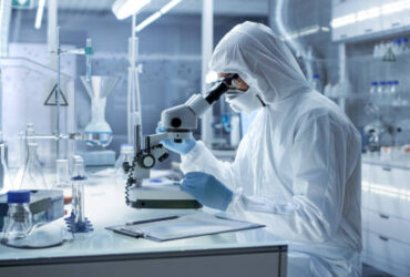 In a Secure High Level Research Laboratory Scientist in a Coverall Examines Petri Dish Under Microscope.
